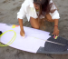 Woman waxing a light purple surfboard on the sand.