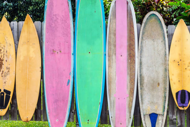 A vibrant row of used, colorful surfboards leans against a weathered wooden fence outdoors.