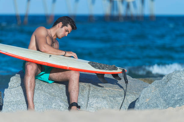 A man sits by the ocean, looking at his surfboard resting on a large rock.