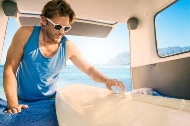 Man in blue tank top waxing surfboard inside van.